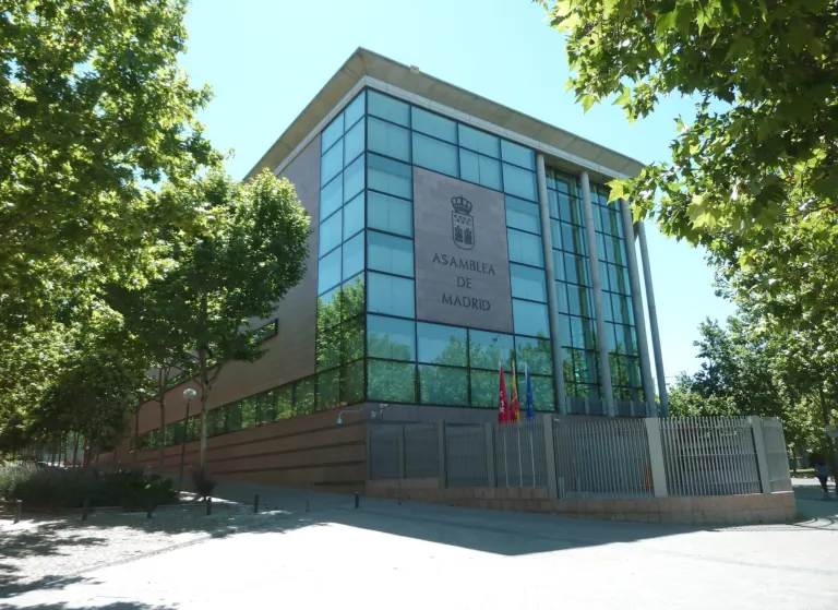 Edificio de la Asamblea de Madrid, con fachada de cristal y placa con el escudo y el nombre, entre árboles y con banderas delante de la entrada