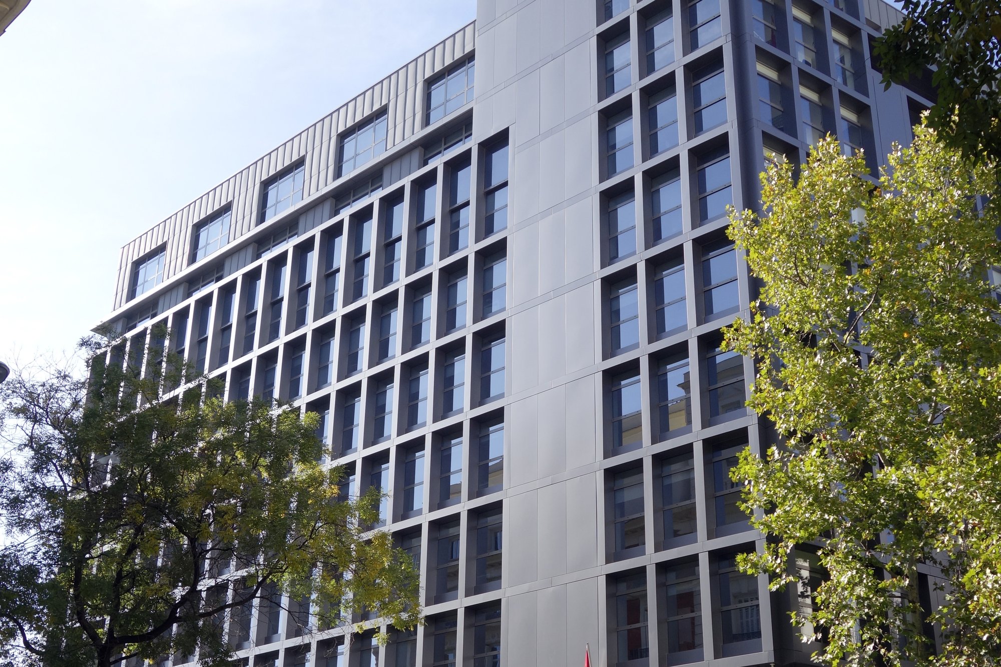 Edificio de oficinas de varias plantas con fachada gris y ventanas en cuadrícula, sede de la Audiencia Nacional, entre árboles