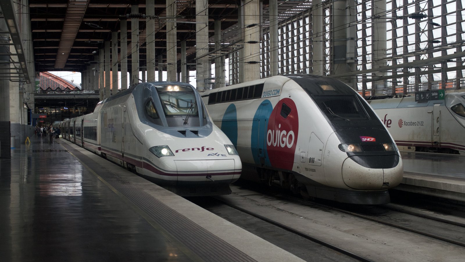 Un tren AVE de Renfe, blanco con franja morada, junto a un tren de Ouigo, blanco y azul, en los andenes cubiertos de la estación de Atocha
