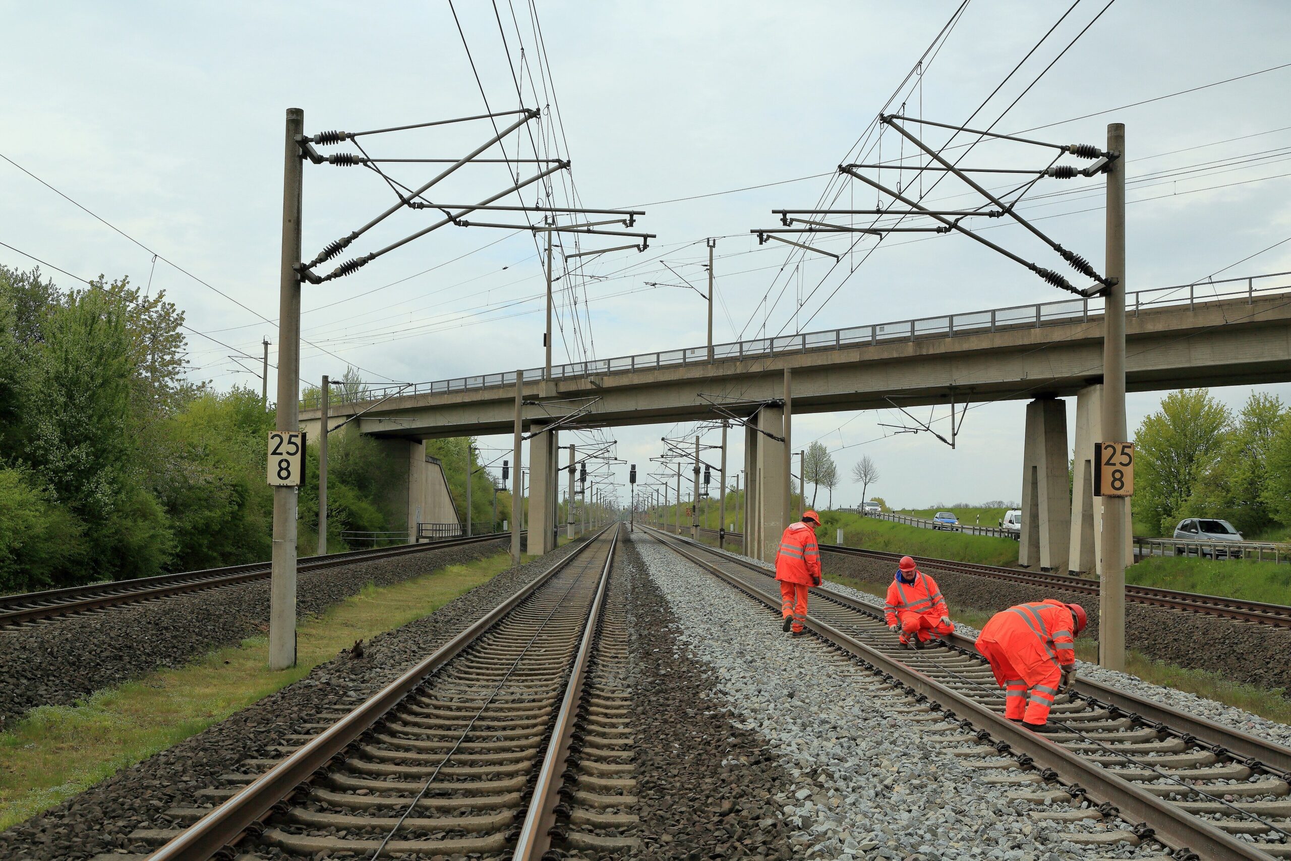 Tres operarios con ropa naranja de trabajo tienden un cable entre los carriles de una vía de tren electrificada, con un puente al fondo