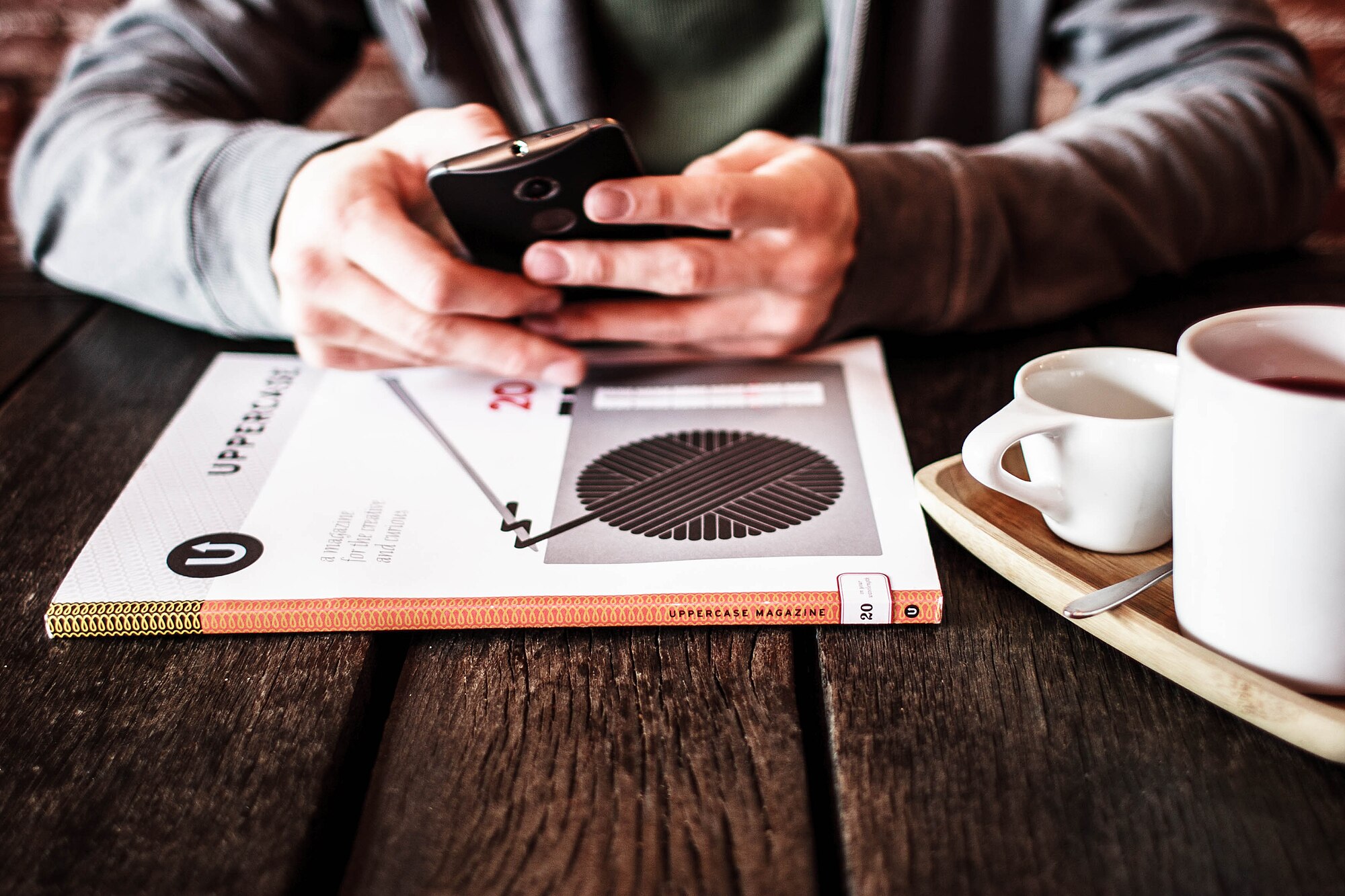 Manos de una persona escribiendo en un teléfono móvil sobre una mesa de madera, junto a una revista y dos tazas de café
