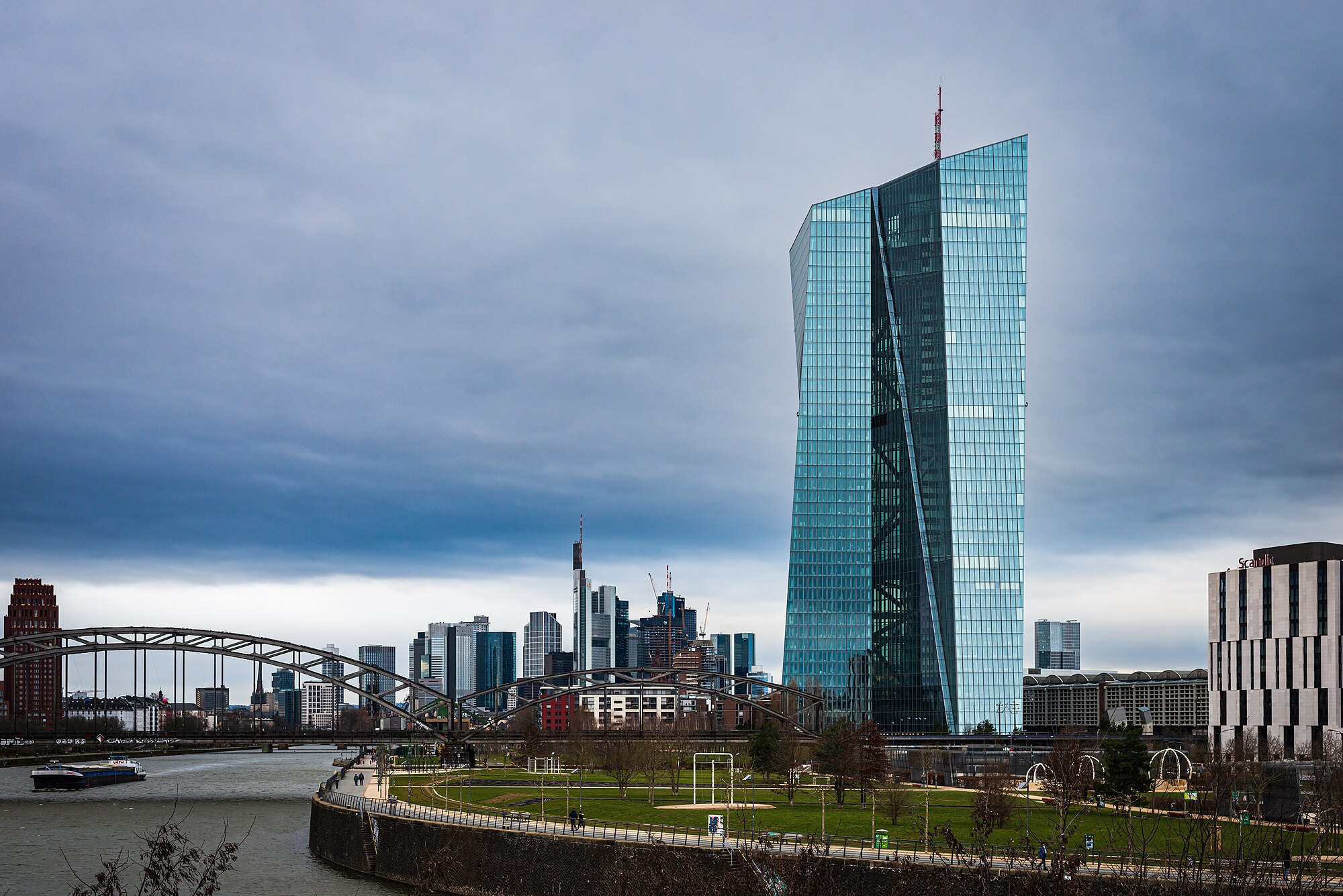 Torre de cristal de la sede del Banco Central Europeo en Fráncfort, junto al río Meno, con los rascacielos de la ciudad al fondo y el cielo nublado