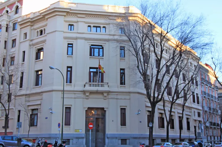 Edificio de esquina de color claro, sede del Centro de Investigaciones Sociológicas, con una bandera de España en el balcón