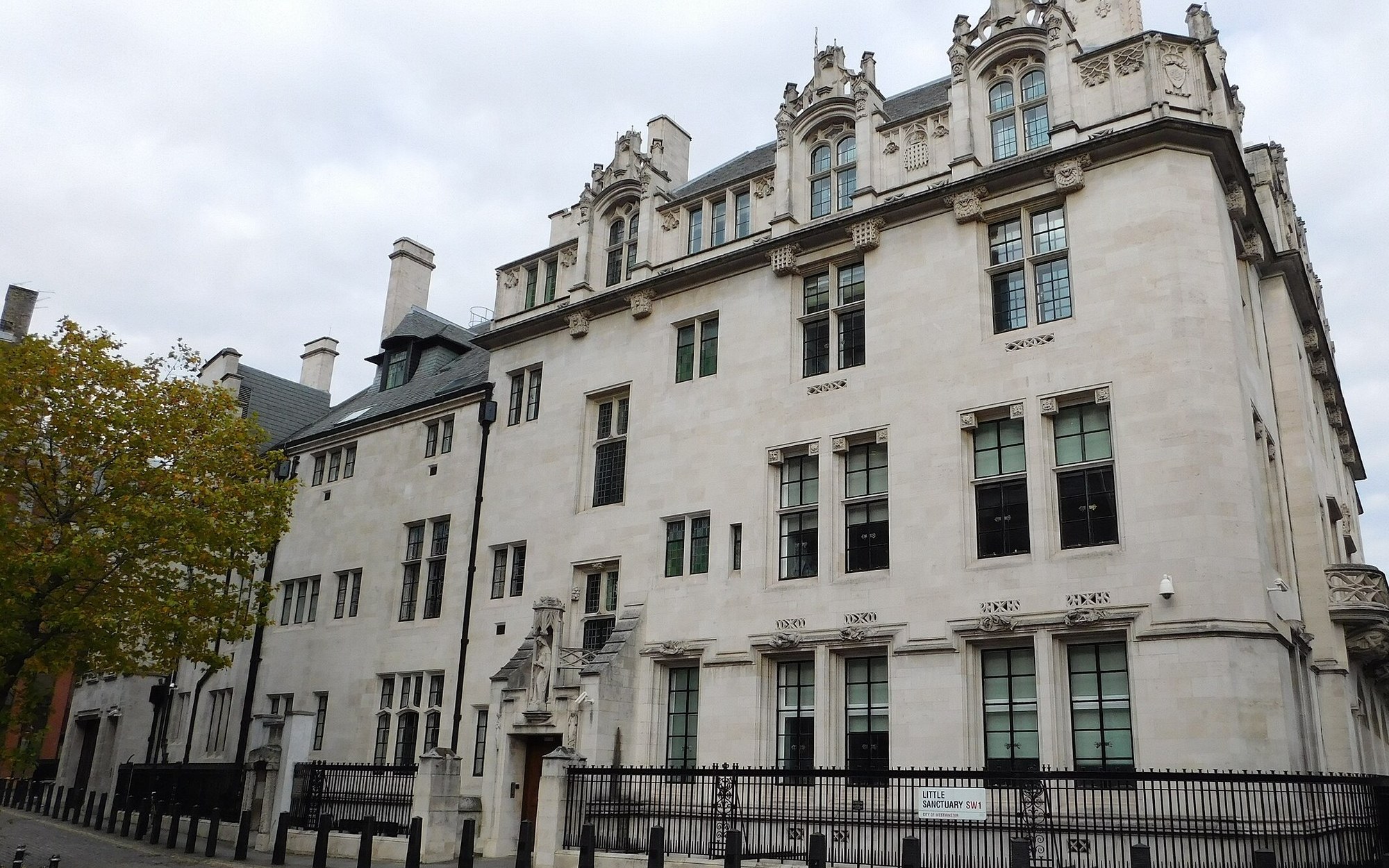 Edificio de piedra clara del Middlesex Guildhall, sede del Tribunal Supremo del Reino Unido en Londres, con una verja negra delante y un árbol a la izquierda