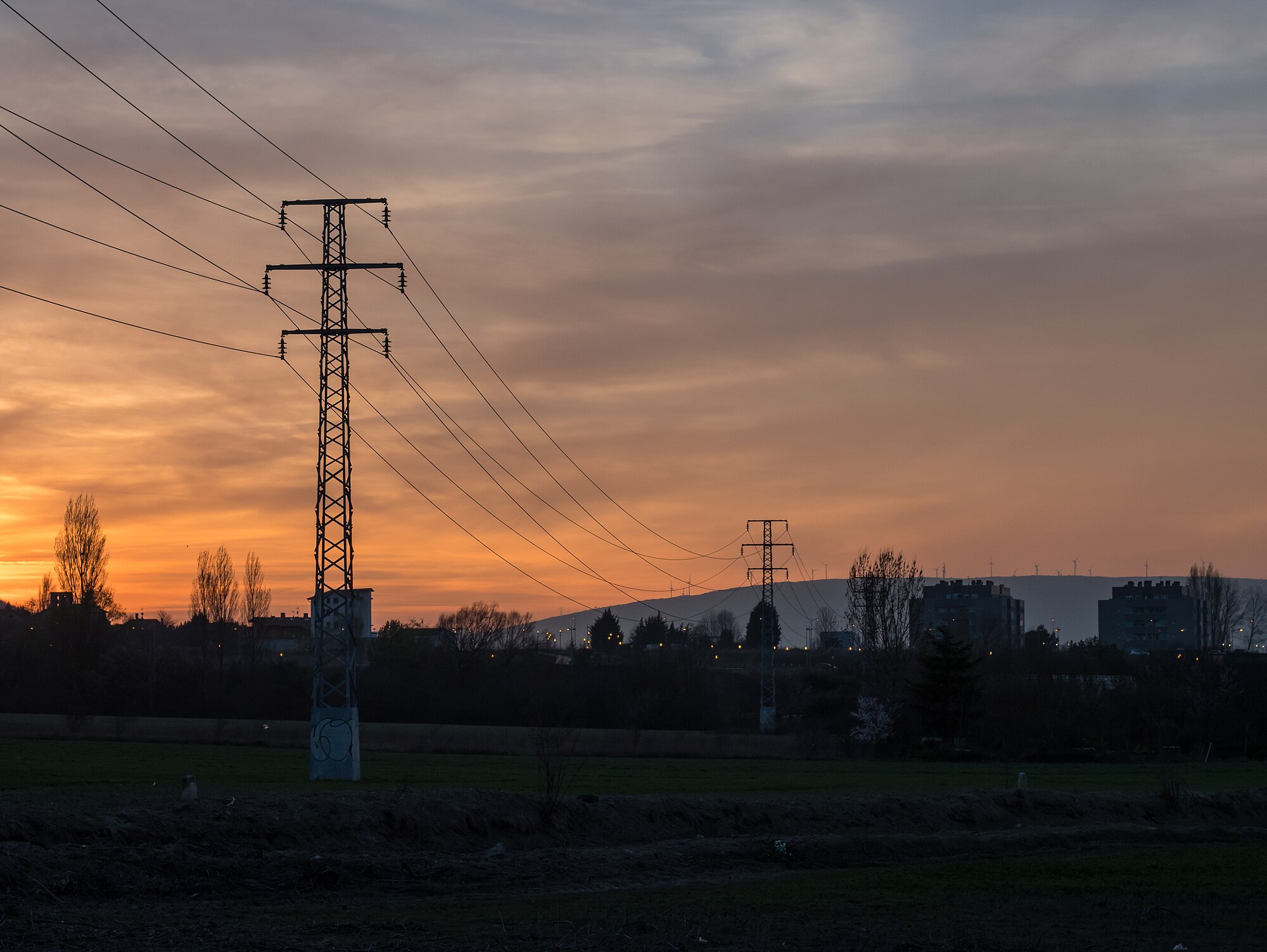 Torres y cables de alta tensión recortados contra un cielo naranja al atardecer, con campos y edificios al fondo