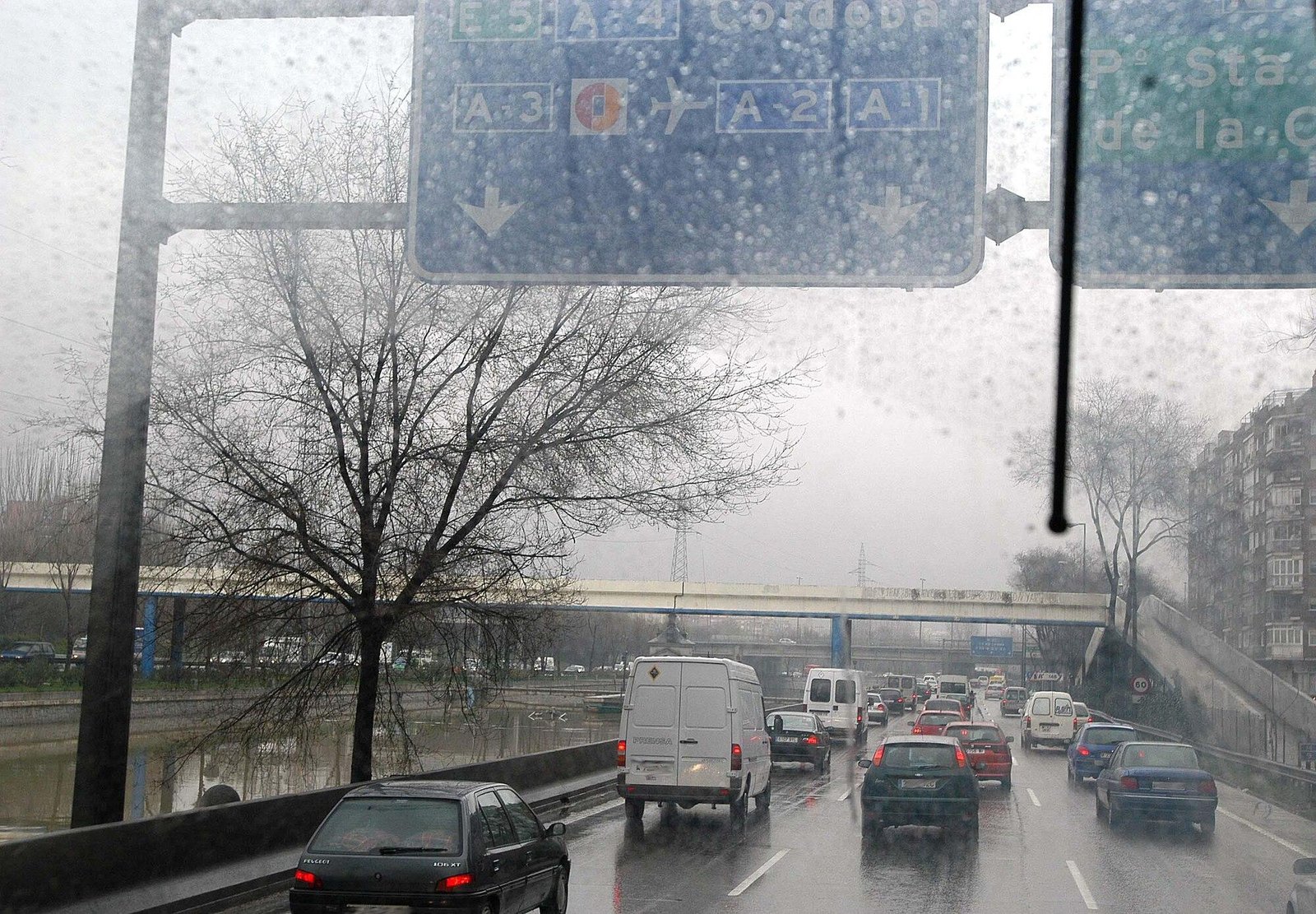 Coches y furgonetas en tráfico lento por una autovía urbana mojada por la lluvia, vistos desde detrás de un parabrisas, con un puente y señales de dirección al fondo