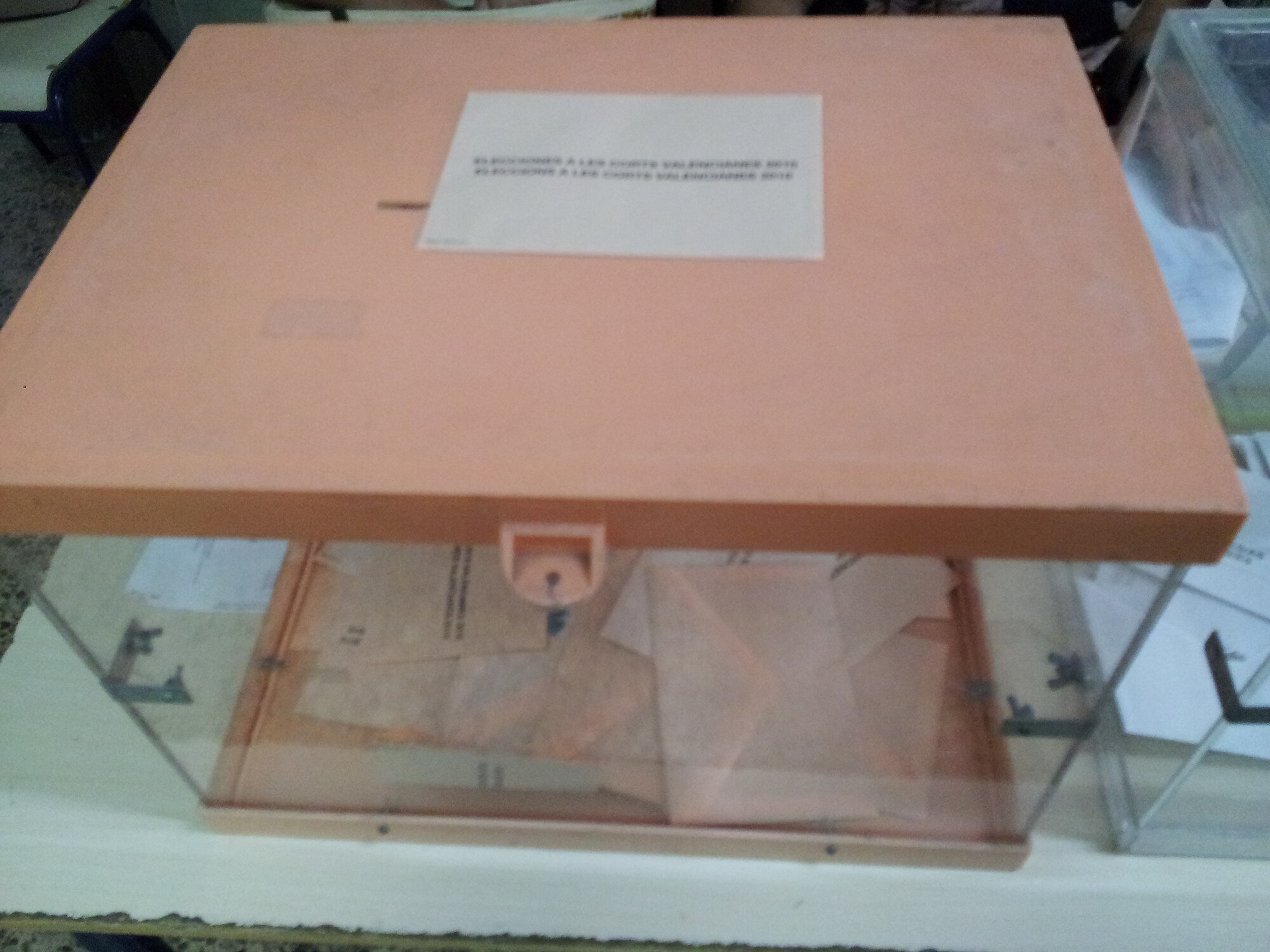 Urna electoral transparente con la tapa de color salmón y sobres de votación dentro, sobre una mesa